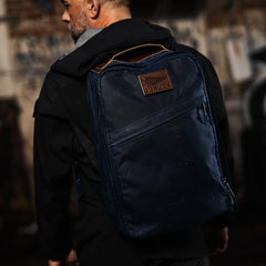Man wearing a black jacket and carrying the GR1 USA - Heritage Waxed Canvas backpack in navy blue, featuring a brown leather patch on the back.
