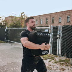 Man lifting GORUCK 100 lb sandbag outdoors, showcasing rucking fitness gear