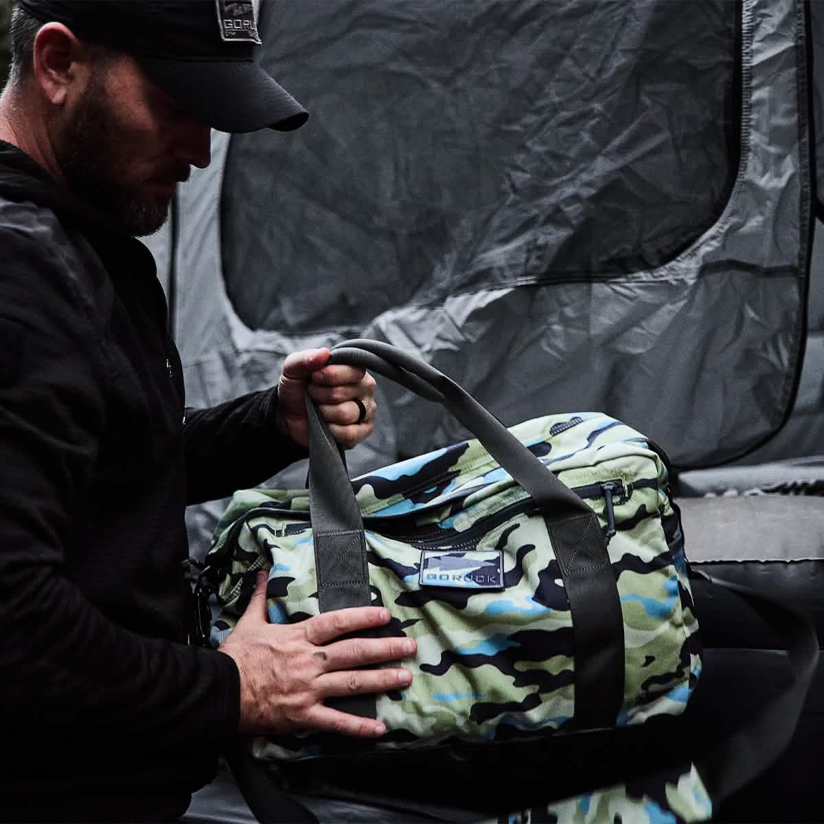 Dressed in a dark outfit, someone tightly holds a GORUCK Kit Bag with robust handles and a convenient shoulder strap, making it ideal for any adventure. Behind them stands an imposing gray tent-like structure, enhancing the atmosphere of travel and exploration.