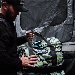Dressed in a dark outfit, someone tightly holds a GORUCK Kit Bag with robust handles and a convenient shoulder strap, making it ideal for any adventure. Behind them stands an imposing gray tent-like structure, enhancing the atmosphere of travel and exploration.