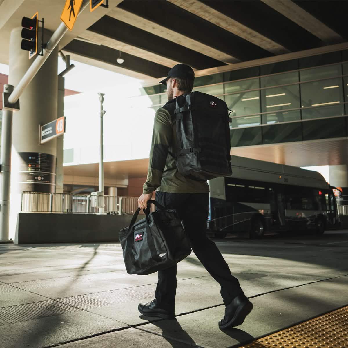 Man carrying GORUCK black backpack and bag outside airport terminal, urban setting