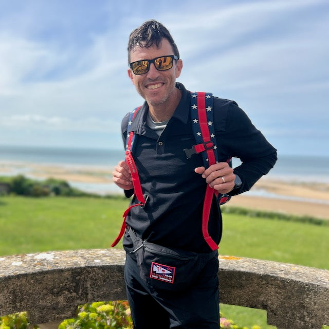 A smiling man in sunglasses with a backpack and the Tactical Fanny Pack - Cordura stands outdoors by the beach on a sunny day.