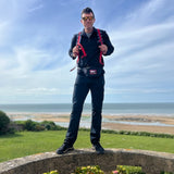 A man in black clothes with a red backpack stands on a stone wall by the beach under a blue sky, wearing Size 34x32 Black only - Men’s Simple Pants - Lightweight ToughDry®—the ideal choice for traveling in style and comfort thanks to their tough, lightweight fabric.