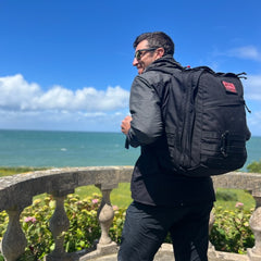 A man with a black backpack stands on a stone balcony, smiling at the ocean under a bright blue sky, wearing Size 34x32 Black only - Men’s Simple Pants made from Lightweight ToughDry® fabric—an ideal choice for those seeking the best travel pants.