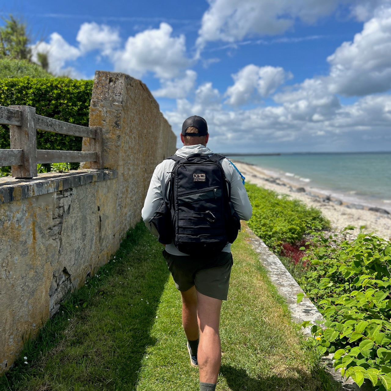 A person carrying the Rucker - Long Range walks a narrow seaside path under a partly cloudy sky, enjoying the benefits of distance rucking.
