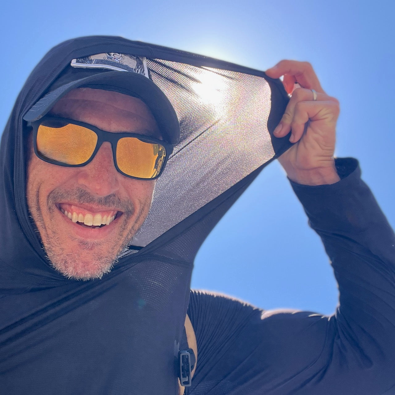 A smiling man in sunglasses and a hat pulls the hood of the Men's Sun Hoodie - ToughMesh over his head, enjoying the sun with UPF 30+ protection beneath a bright blue sky.