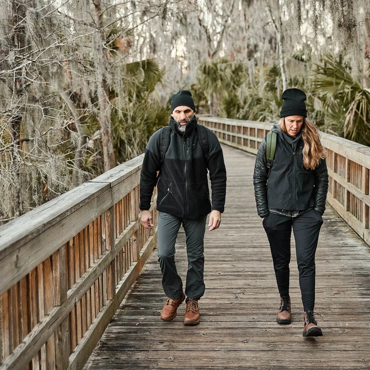 Two people wearing GORUCK gear hiking on a wooden boardwalk in a forested area