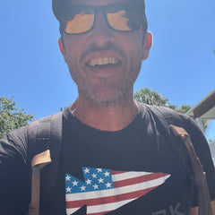Man smiling outdoors in bright sunlight, wearing sunglasses, a cap, a U.S. flag arrow shirt, and the Ruck Plate Carrier 3.0 backpack—ready for his rucking workout.