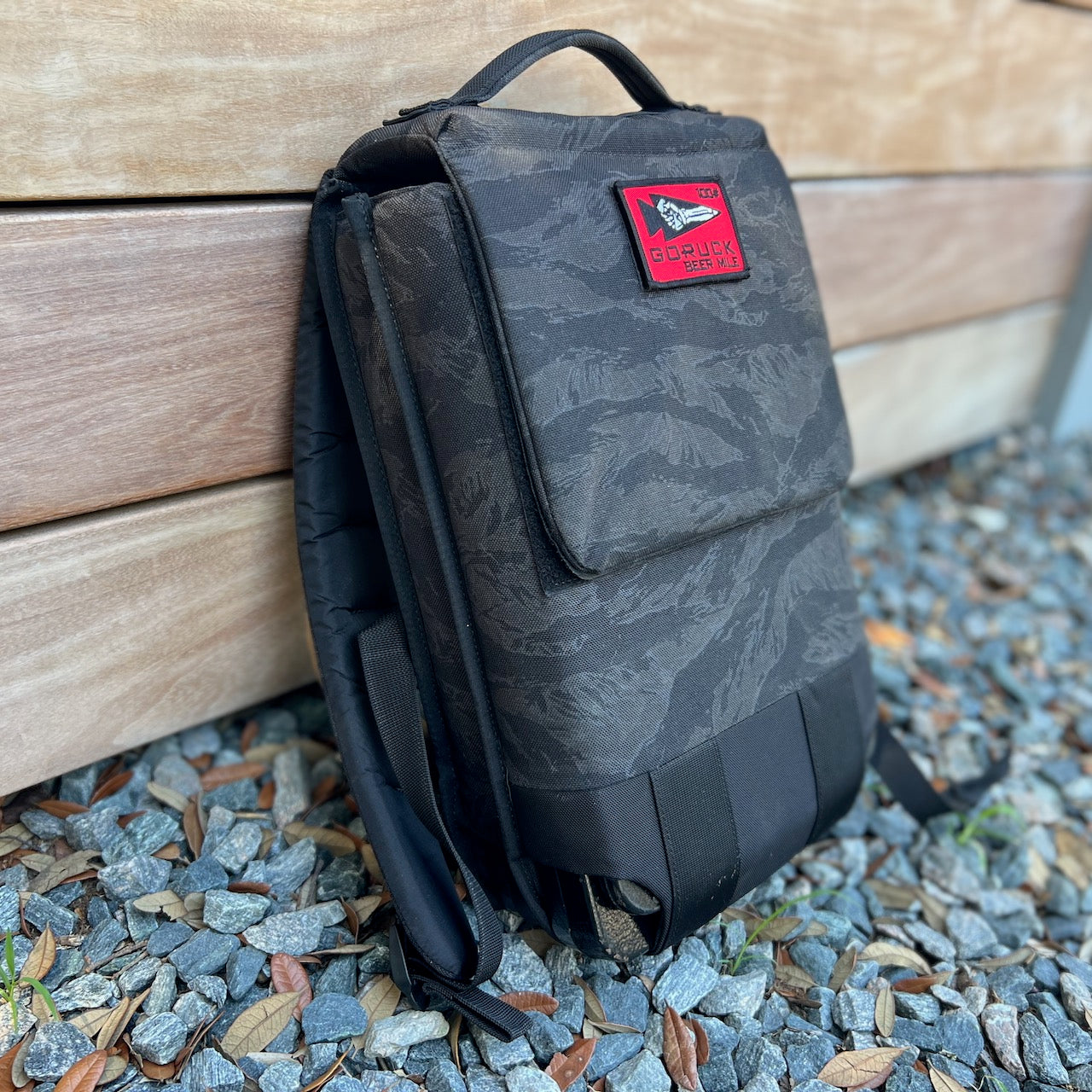 The Ruck Plate Carrier 3.0, featuring black camo with a red patch, is perfect for rucking workouts and is shown leaning against a wooden wall on gravel and leaves.