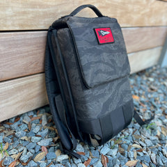 The Ruck Plate Carrier 3.0, featuring black camo with a red patch, is perfect for rucking workouts and is shown leaning against a wooden wall on gravel and leaves.