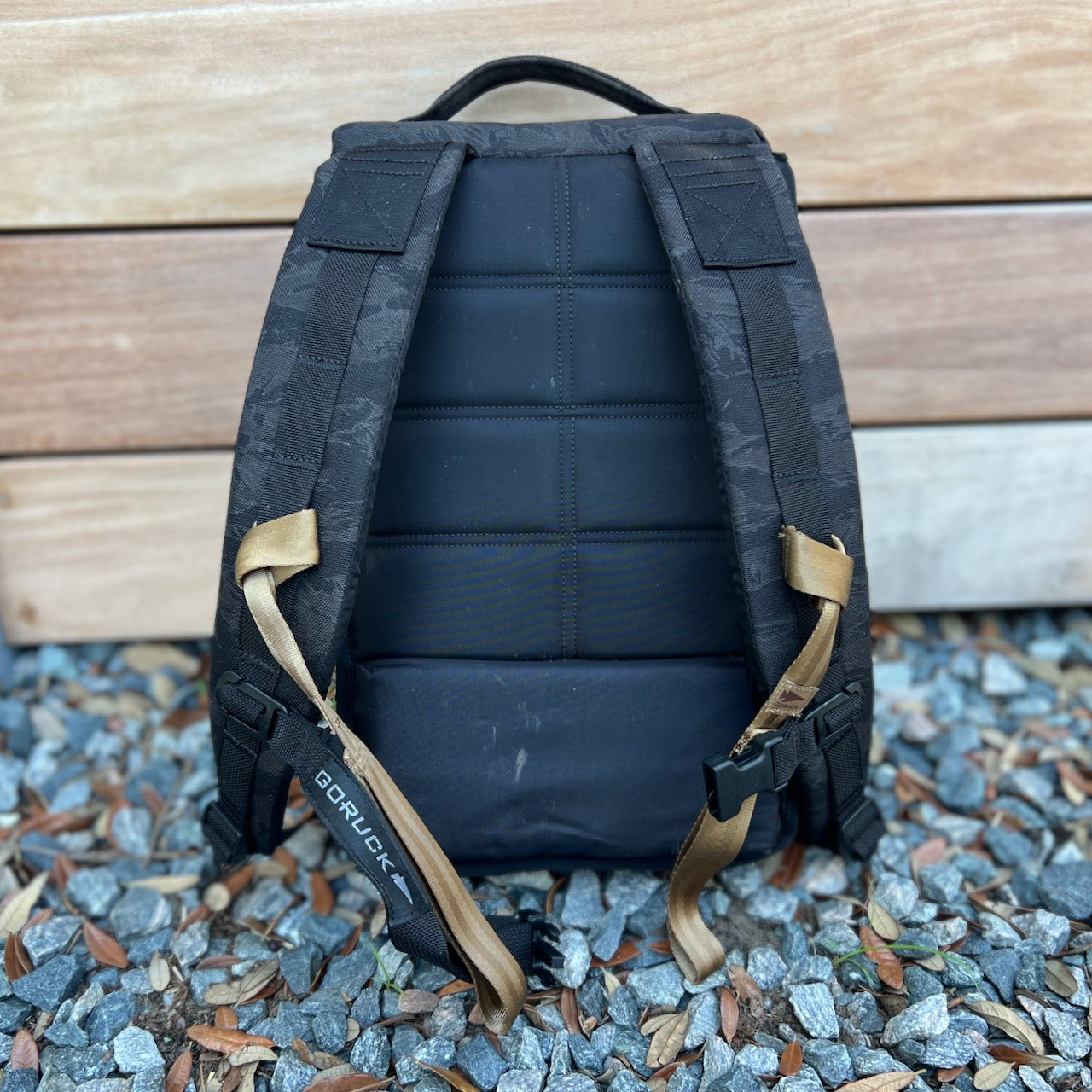 The Ruck Plate Carrier 3.0, a black backpack with padded straps and tan buckles, rests on gravel near wooden planks—ideal for rucking workouts or use as a versatile plate carrier.