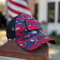 The Cadre Hat - CORDURA®, featuring a red, white, and blue camouflage pattern with a patch, is crafted from 500D CORDURA® and military-grade mesh, displayed in front of a blurred American flag and building.