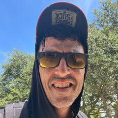 A man in sunglasses and a hoodie smiles outdoors, wearing a Cadre Hat – CORDURA®, crafted from rugged 500D CORDURA, with trees and a blue sky in the background.