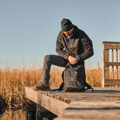 Man in outdoor gear kneeling on wooden dock with GORUCK rucksack in marshland setting