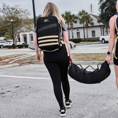 Woman rucking outdoors with GORUCK backpack and sandbag, demonstrating tough rucking gear.