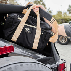 GORUCK black duffel bag with beige straps being loaded into a car trunk outdoors