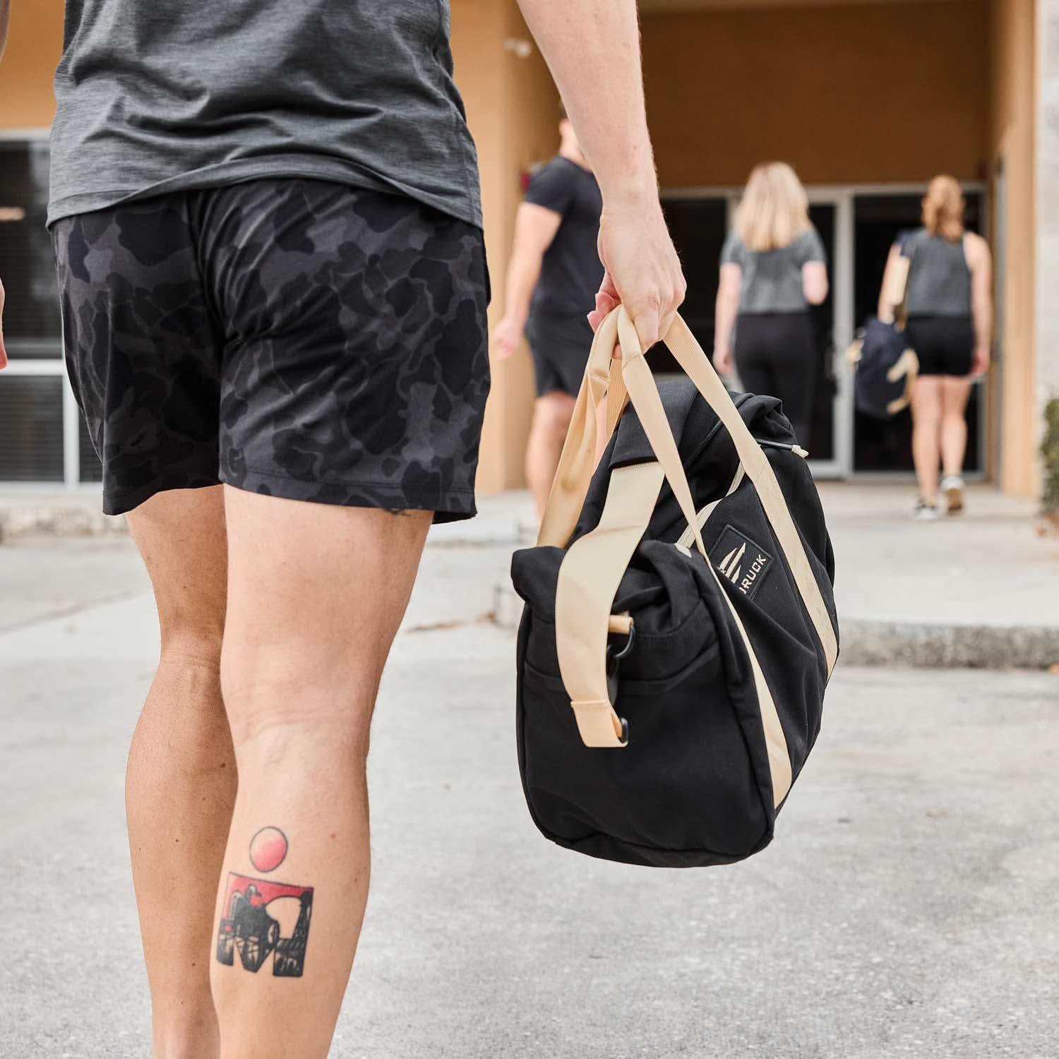 Person carrying GORUCK gym duffel bag, walking toward building with rucking group outside