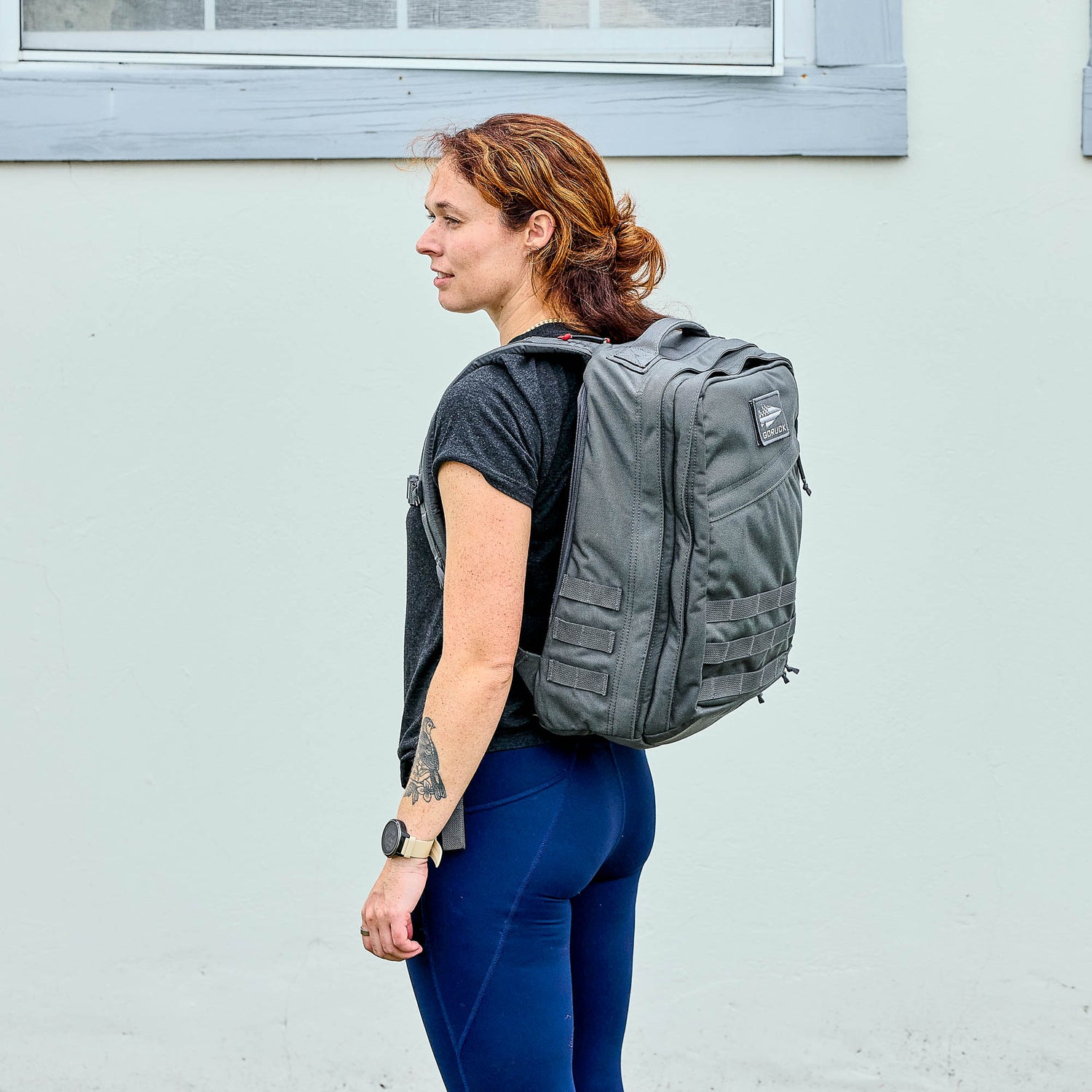 A woman with a tattoo stands sideways against a light-colored wall, wearing the GR2 - Cordura gray travel backpack.