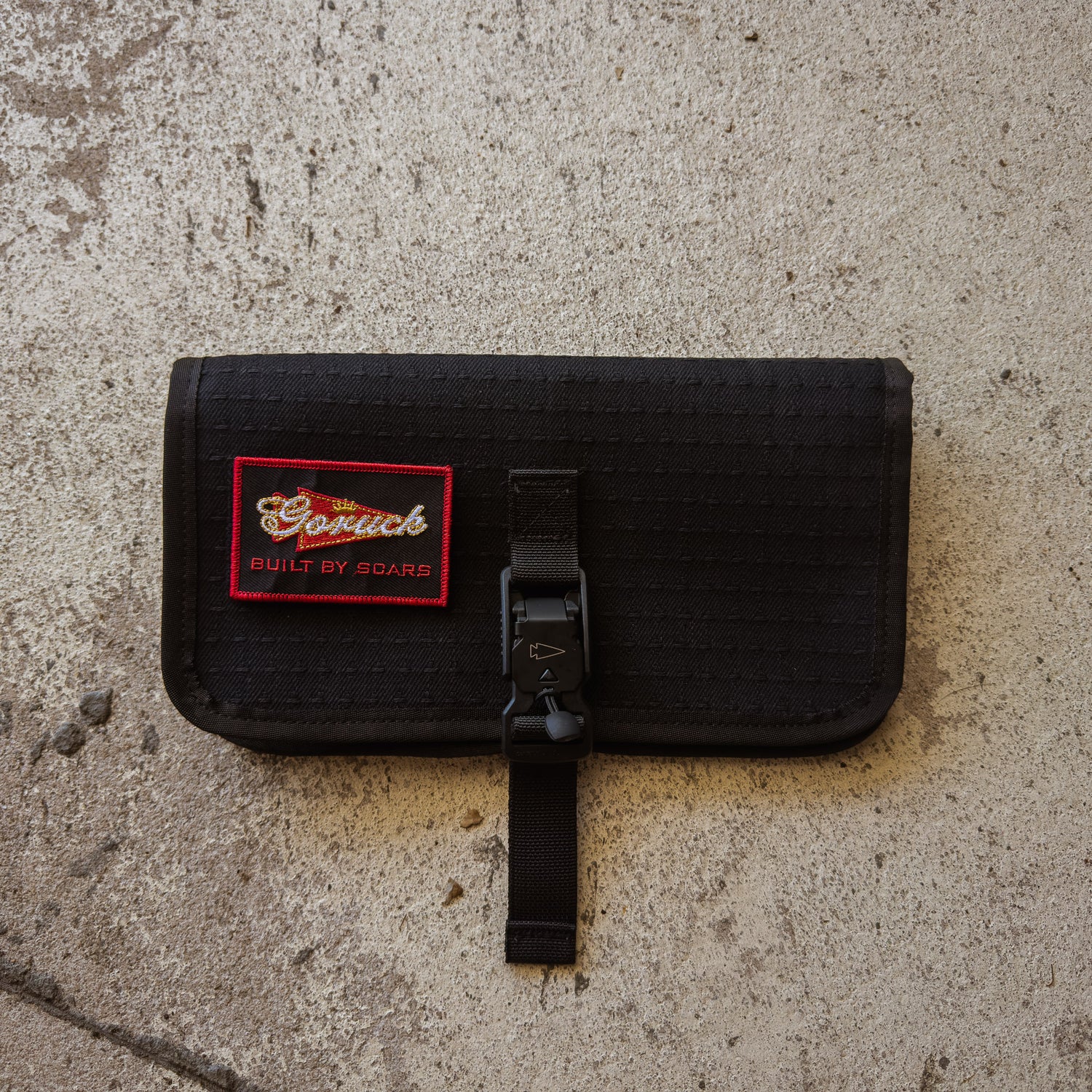 The Scars Wire Dopp, a black waxed twill EDC pouch with a red patch and buckle, rests on a textured concrete surface.