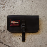 The Scars Wire Dopp, a black waxed twill EDC pouch with a red patch and buckle, rests on a textured concrete surface.
