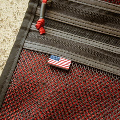 The Scars Wire Dopp is a red and black mesh travel organizer with zippers, red cord pulls, and a small American flag tag.