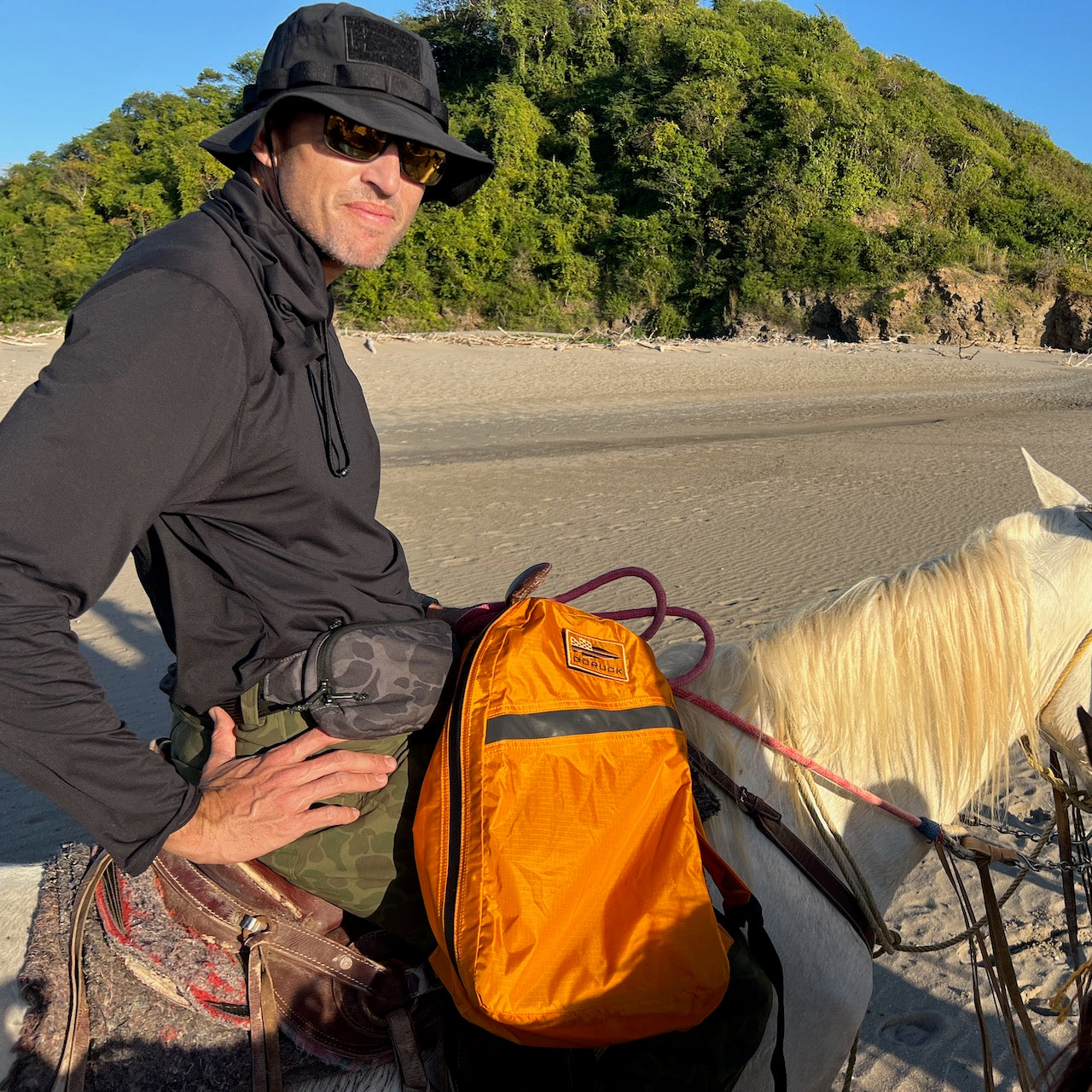 A man wearing the Boonie Hat - Tactical - ToughDry and sunglasses rides a white horse along a sandy beach with green hills in the background.