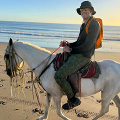 A person wearing the Boonie Hat - Tactical - ToughDry and sunglasses rides a white horse along a sandy beach by the ocean at sunset.
