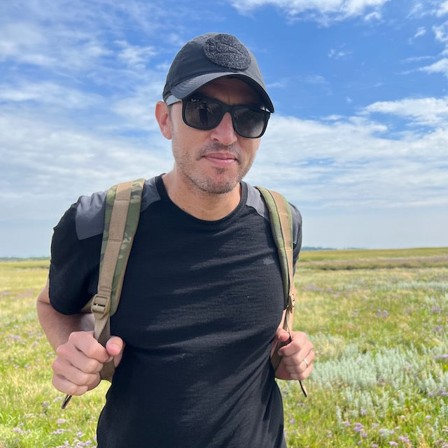 A man in sunglasses and a cap, wearing the Men’s Commando Tee - Merino Wool and carrying a backpack, stands in a grassy field under a blue sky.