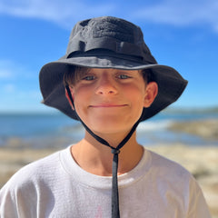 Smiling boy in a white shirt wearing the Boonie Hat - Tactical - ToughDry, standing outdoors by the beach on a sunny day.