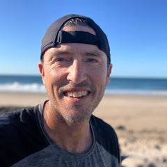 Smiling man in a Performance Running Hat - Italian ToughMesh at a sunny beach with ocean waves and a blue sky in the background.