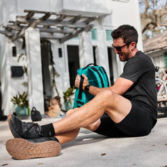 Man in black shirt and shorts laughs while sitting on pavement, holding a teal bag with a house behind—wearing Merino Challenge Socks - Trail Cuff for ultimate comfort on the go.