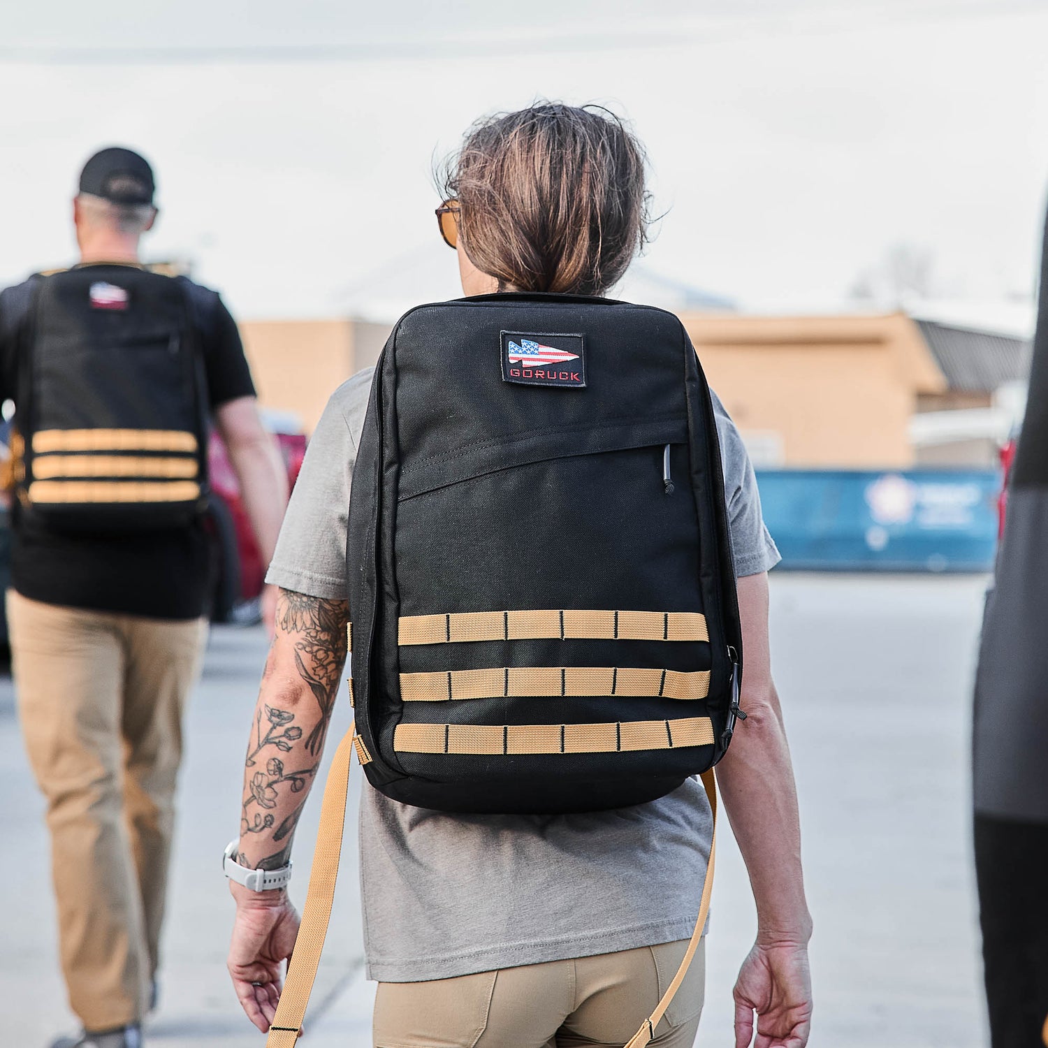 A person with a tattooed arm is wearing a black GR1 rucksack while walking outdoors with others dressed similarly.