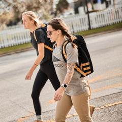 Two women with GR1 backpacks, made from Special Forces grade materials, walk across a sunny street in athletic wear.