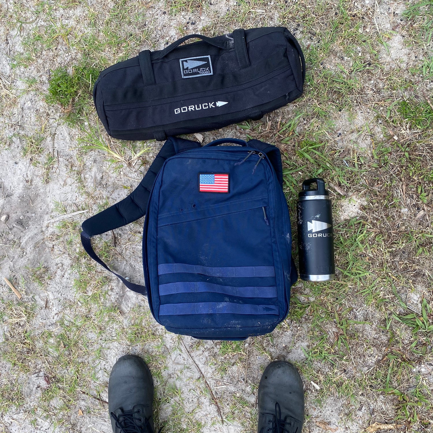 A pair of boots, a black GORUCK bag, a US flag-patched navy backpack, and Simple Training Sandbags with a GORUCK bottle sit on the grass—ideal for sandbag training.