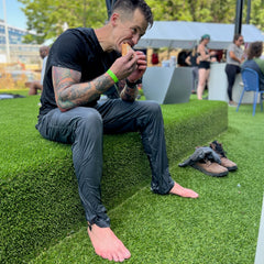A man in wet clothes sits barefoot on artificial grass, eating a sandwich with his MACV-2 - Mid Top - Chestnut + Black boots and gloves beside him, the aggressive outsole of his Special Forces boots visible.