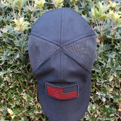 A Cadre Hat - CORDURA®, featuring a black military-grade design and a red patch, rests atop green, spiky holly leaves.