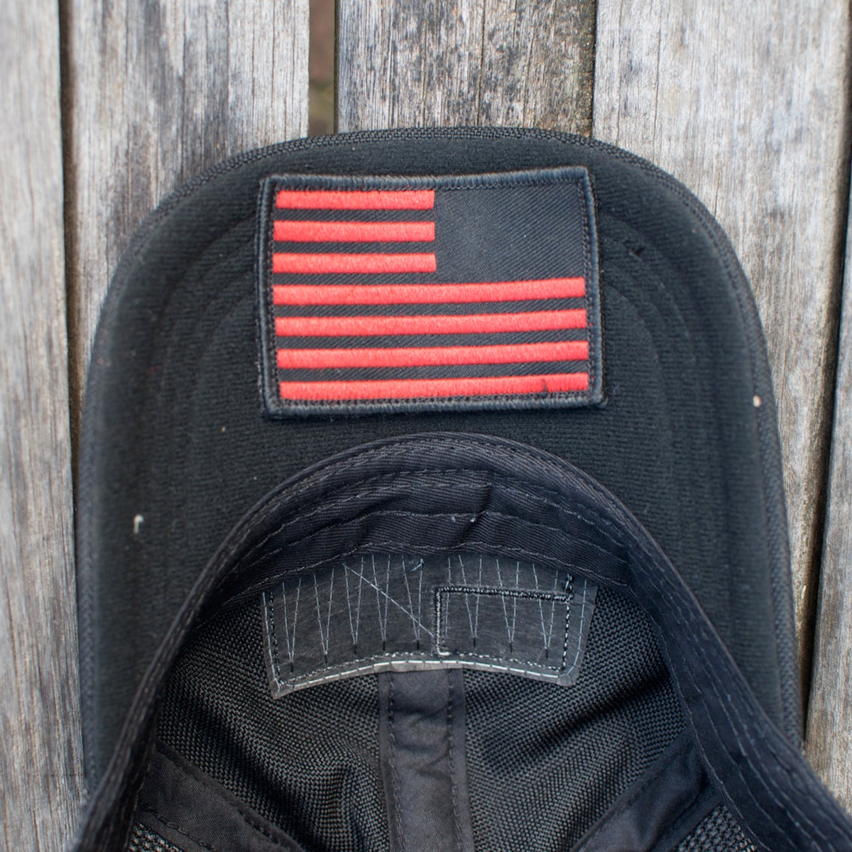 The Cadre Hat - CORDURA® is a black, military-grade cap featuring a red, stylized American flag patch. It’s crafted from rugged 500D CORDURA® fabric and shown on a weathered wooden surface.