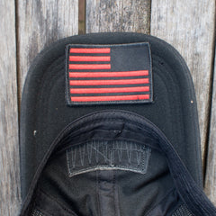The Cadre Hat - CORDURA® is a black, military-grade cap featuring a red, stylized American flag patch. It’s crafted from rugged 500D CORDURA® fabric and shown on a weathered wooden surface.