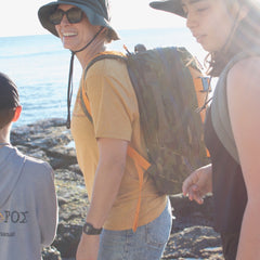 Three people, each with hats and backpacks—one carrying the Double Bullet Ruck 18L featuring a bombproof laptop compartment—walk along a rocky beach in bright sunlight.