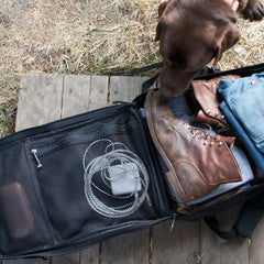 A brown dog sniffs a GR2 USA - Cordura carry-on bag with boots, jeans, and cables inside on a wooden deck.
