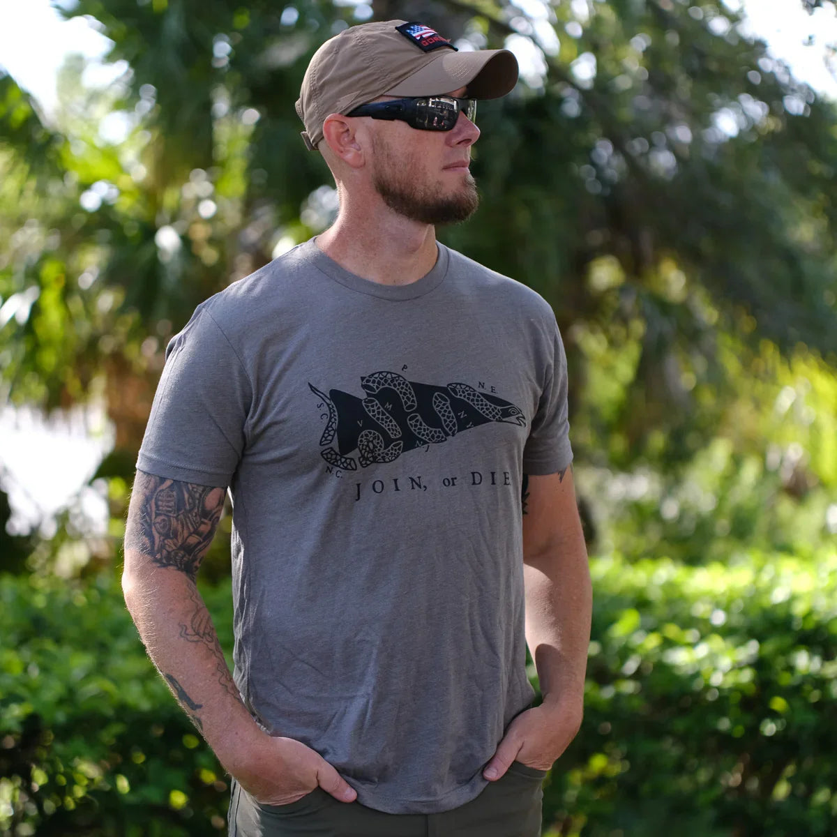 Man outdoors wearing GORUCK Join or Die grey graphic t-shirt, tan cap, and sunglasses