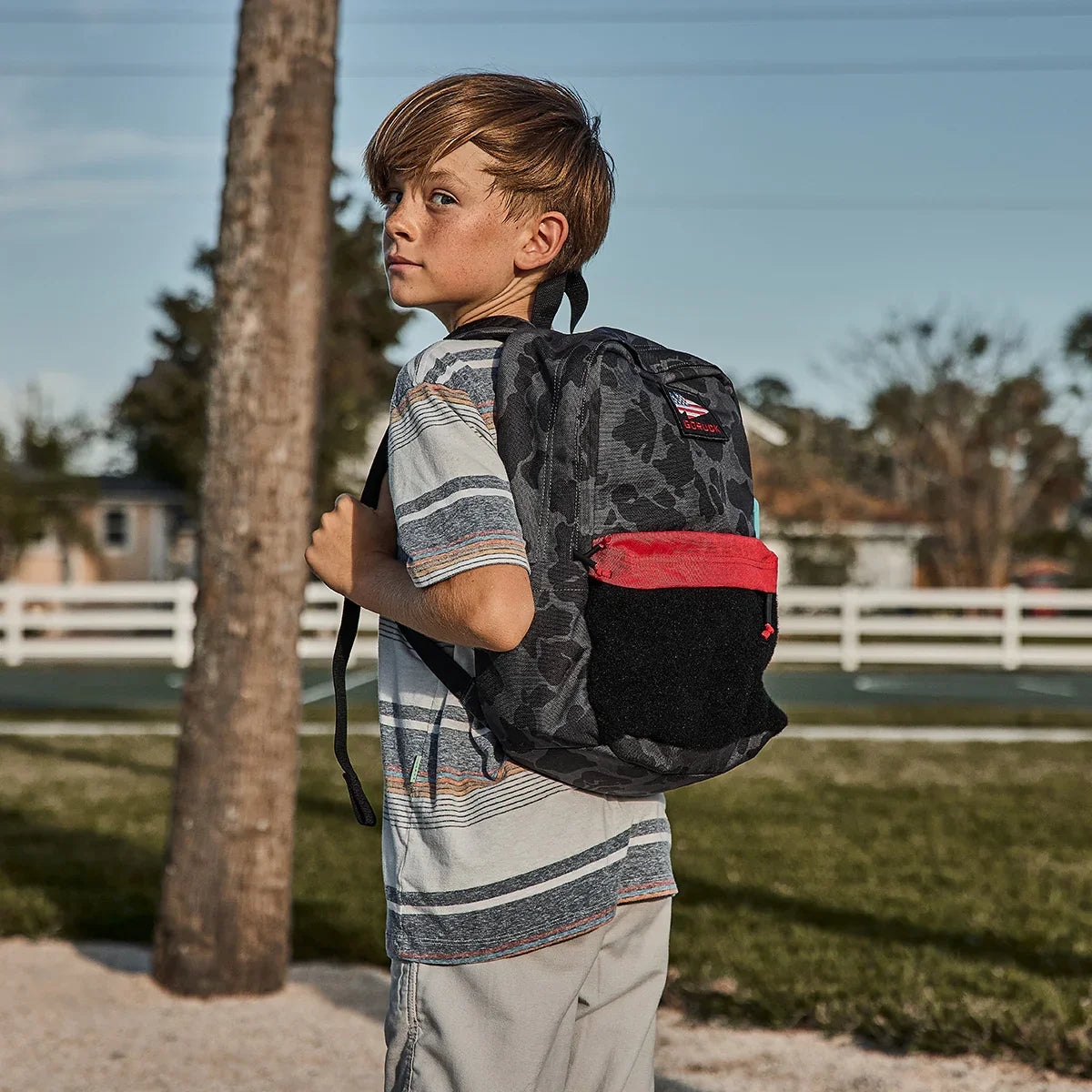 Boy wearing a GORUCK Kid Ruck backpack outdoors, featuring black camo and red pocket.