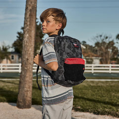 Boy wearing a GORUCK Kid Ruck backpack outdoors, featuring black camo and red pocket.