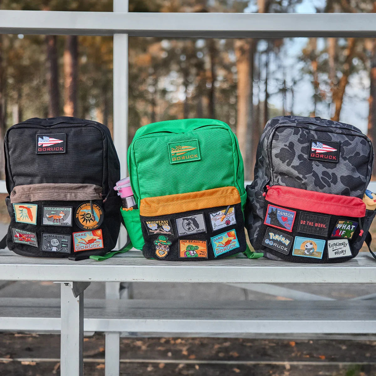 Three GORUCK backpacks for kids with colorful ruck patches displayed outdoors on a bench.