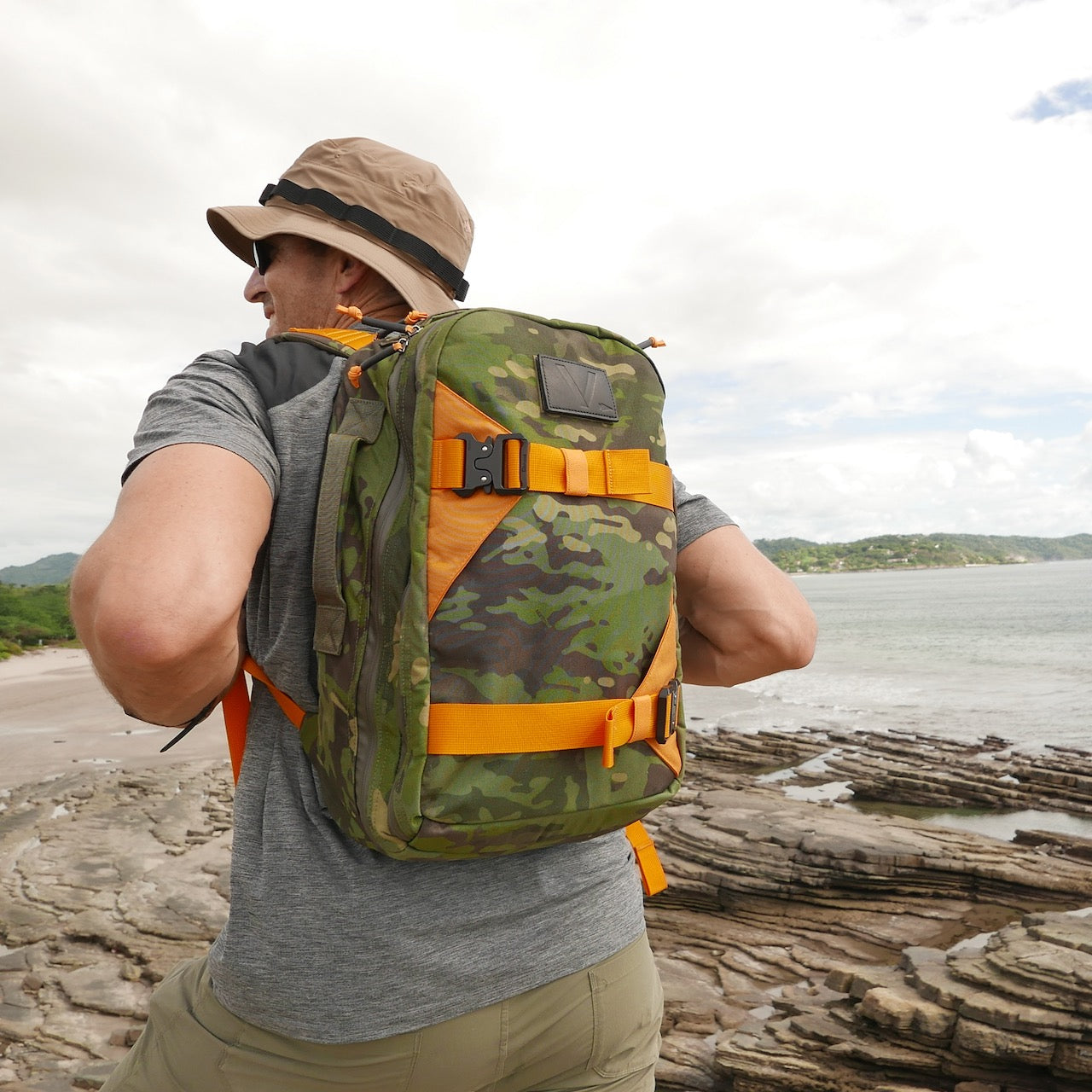 A person stands on a rocky coastline by the sea, wearing a hat and carrying the Double Bullet Ruck 18L with Bombproof Laptop Compartment.
