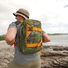 A person stands on a rocky coastline by the sea, wearing a hat and carrying the Double Bullet Ruck 18L with Bombproof Laptop Compartment.
