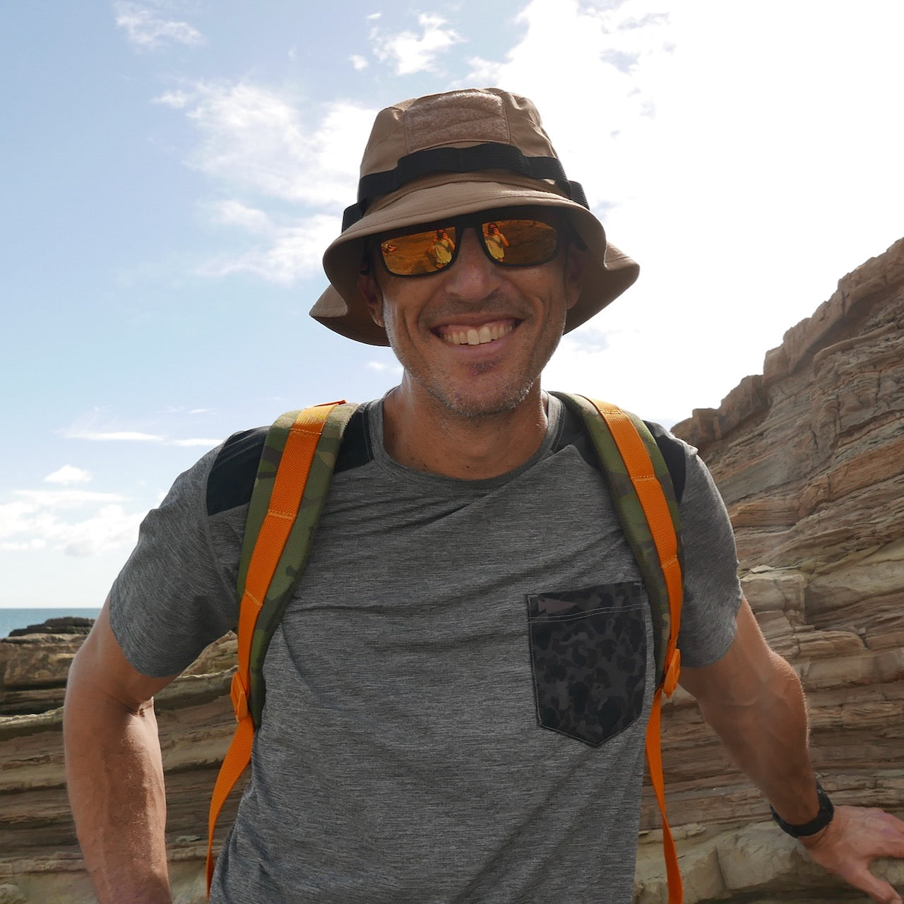 Man wearing the Boonie Hat - Tactical - ToughDry and sunglasses, smiling in a grey ToughDry shirt with a backpack, standing by rocky cliffs outdoors.