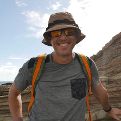 Man wearing the Boonie Hat - Tactical - ToughDry and sunglasses, smiling in a grey ToughDry shirt with a backpack, standing by rocky cliffs outdoors.