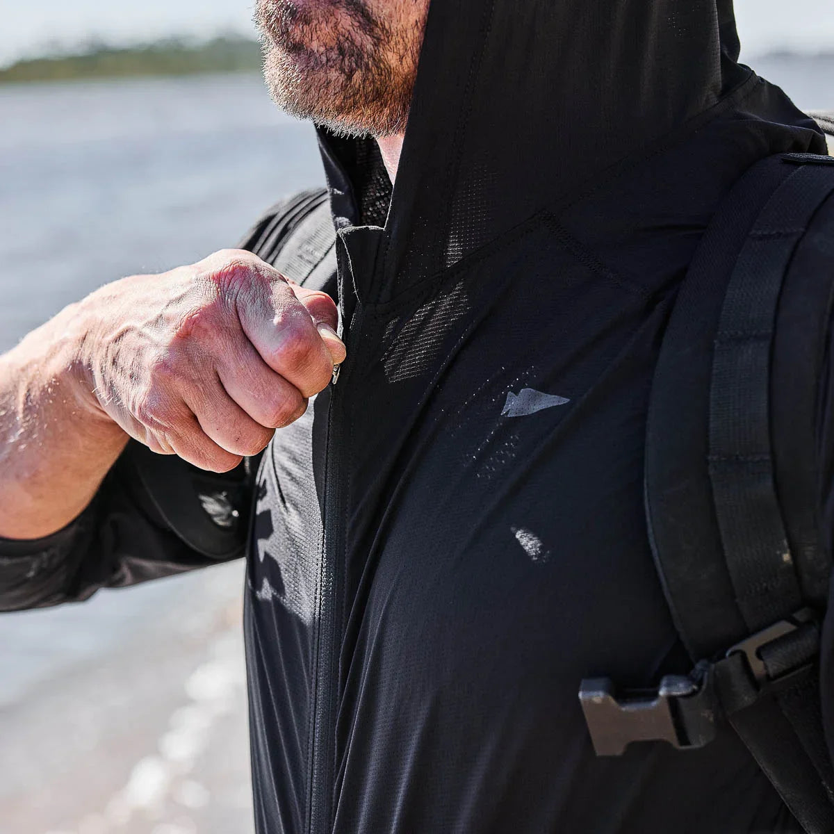 Man zipping up a black GORUCK performance longsleeve hoodie by the water