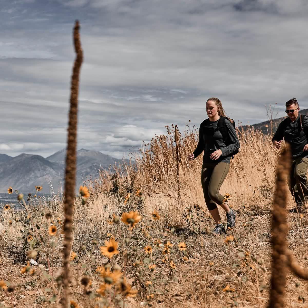 Two people rucking outdoors on a mountain trail with backpacks, wildflowers, and dry grass.